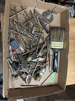 Photo showing a cardboard box filled with an assortment of various used metal drill bits of different sizes, a large paintbrush with black handle and natural bristles, grinding disc, wire brushes and various other small hand tools and metal parts.