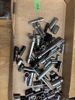 Assorted used metal sockets and extension bars spread loosely in a cardboard box.