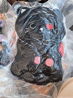 Small black cat squishie plush toy vacuum packed in clear plastic bag showing stitched scar, heart patch on eye, and red patches.