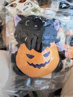 Small black squishie cat holding orange pumpkin with jack-o-lantern face in clear vacuum sealed packaging