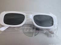 Photo showing front and side view of white frame rectangular sunglasses with black lenses sealed in plastic wrapping.