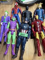 Photo showing seven various comic action figures laid out on a wooden surface including Spider-Man, Black Panther, Iron Man, Captain America, Joker, and two others.