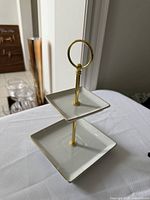 Front view of two-tier dessert tray showing white square plates with gold trim and gold handle ring