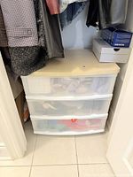 Photo of white framed plastic storage organizer with three wide translucent drawers, wood-colored top, located in a closet space with clothing around it.
