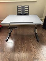 Front view of the black adjustable lap desk with visible legs and book holder on hardwood floor.