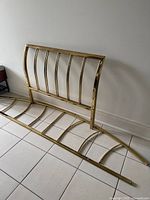 Disassembled antique gold metal daybed frame parts leaning against a wall on tiled floor.