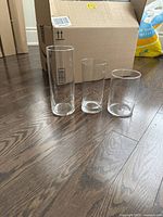 Three clear glass cylinder vases in small, medium, and large sizes arranged on wood floor in front of a cardboard box.