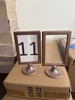Two bronze-coloured small rectangular frames with stands, one contains a paper insert with the number 11 and the other is empty, both standing on a cardboard box.