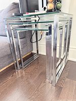Three glass nesting tables with silver metal frames nested together on brown wooden floor next to gray sofa.