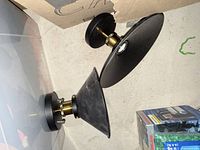 Photo showing two industrial style light fixtures placed on a floor side by side. One fixture is smaller with a narrow black metal shade and brass fittings, the other is larger with a wider black metal shade and brass fittings.