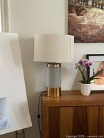 Side view of one table lamp on wooden cabinet, showing gold metal base and white fabric shade, cord visible.