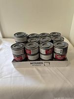 Multiple views of 12 cans of methanol gel chafing fuel packed in a cardboard tray showing unopened cans with metal lids and labels with safety and usage information.