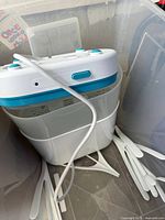 Sterilizer unit with power cord and various white plastic accessories like tongs or spatulas placed inside a transparent container.