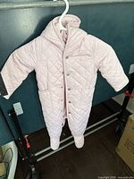 Front view of pink quilted snowsuit hanging on a hanger showing full body coverage, snap buttons, and pocket details.