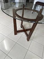 Side angle of round glass dining table showing clear thick glass top and dark stained wood base with circular frame and cross supports.