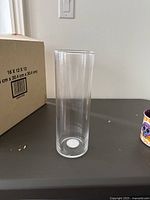 Single tall clear glass cylinder vase on black surface with light switch in background.