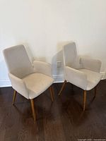Two light grey upholstered dining chairs with armrests and wooden legs positioned against a white wall on dark hardwood floor.