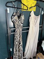 Two women’s evening gowns hanging on black hangers on a black rack, showing full length and detail of the zebra print one-shoulder dress and ivory/cream dress.