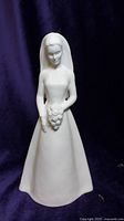 Front-facing photo of the all-white bone china figurine depicting a bride with bouquet and veil, set against dark purple background.