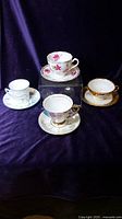Four fine china teacups and saucers with floral and gold designs on dark background.