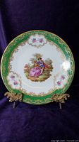 Front view of the porcelain plate showing a hand-painted Victorian scene of a couple in period dress with floral and gold pattern border on green background.