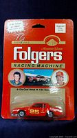 Front view of the sealed Ertl 1/64 scale die-cast 25 Folgers Racing Machine in package with red backing card featuring driver images and Folgers logo.