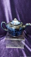Front view of indigo marbled ceramic teapot with lid on a clear display stand against a purple background.