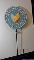 Full front view showing circular blue sign with chicken illustration and text mounted on black metal stake.