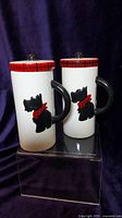 Front view of two tall ceramic mugs each decorated with a black Scottie dog wearing a red plaid scarf, black handles, and red plaid trimmed lids with black knobs.