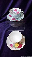 Top view showing two bone china teacup and saucer sets with floral rose patterns, one with red roses and the other with mixed red and yellow roses, placed on clear acrylic stands against a purple velvet background.
