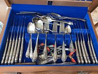 Top view inside the open wooden box showing arranged silverware including forks, spoons, and cutlery pieces with detailed ornate handles in the Oneida Enchantment pattern.