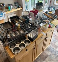 Overall view of baking supply lot on a wooden kitchen island showing muffin pans, tart pans, baking trays, measuring cups, flour sifter, spatulas, and other baking tools.