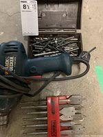 Black & Decker drill on ground next to metal box filled with drill bits and red plastic holder with chisel bits.