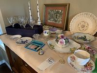 Photo shows tabletop with 8 crystal wine glasses in navy box, framed art, bone china flower bouquets, plates with floral and fruit designs, a match box, and decorative items including a mini fork set.