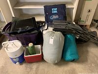Overview of camping lot including two sleeping bags in sacks, foldable wheeled outdoor chair, two tarps, and coolers.