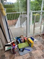 Wide shot of household cleaning supplies arranged near a sliding glass door, showing mops, step stools, and various cleaning bottles.
