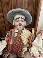 Porcelain clown doll with sad expression, large light blue hat, red plaid tie, and plaid shoulder cloth, showing some wear.