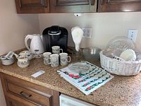 Photo showing Keurig coffee maker, electric kettle, mugs, small dishes, linens, and basket with glass plates on countertop