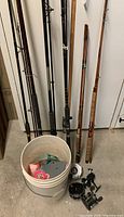 Photo shows six fishing rods leaning against a wall with a white bucket in front containing various fishing accessories.