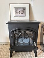 Front view of compact black electric fireplace heater with framed glass door and metal grid design, standing on four legs with carpeted floor.