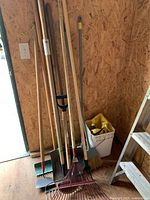 Photo showing the full set of garden tools leaning against a wooden wall including various rakes, shovels, hoes, and brooms.