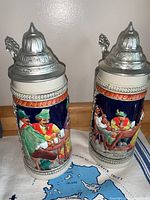 Two vintage German beer steins with colorful raised relief scenes of Bavarian men and pewter lids.