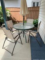 Outdoor patio set with round glass table, beige umbrella, and four folding chairs on a concrete patio near a wooden fence and plants.