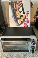 Photo shows a Black+Decker toaster oven in front of a Hamilton Beach griddle box. The box displays breakfast items like pancakes, eggs, bacon, and sausages cooking on the griddle's surface.