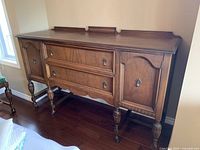 Front and side views of warm wood tone sideboard with two cabinet doors, two drawers, carved wooden legs on caster wheels.