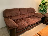 Brown fabric sofa with plush cushions, curved base detail, and padded armrests, shown in a side angle with carpeted floor and wall background.