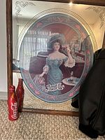 Vintage Pepsi glass mirrored framed image with woman in period attire, with two 2010 Olympics Coca-Cola aluminum bottles in front.