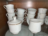 Photo showing stacked white tea cups with gold rims, cream and sugar bowl with lid and creamer pitcher.