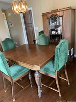 Dining room setup showing vintage wooden dining table with six chairs upholstered in royal green velvet. One chair has armrests (captains chair). Wood cabinet and chandelier in background.