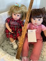 Two reborn dolls seated together with a ruler for size comparison showing approx 14-15 inches tall, one in a red tartan dress with blonde hair, the other in pink overalls with brunette hair in braids.